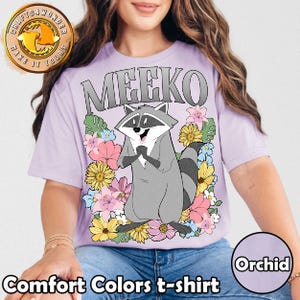 Pu&ograve; includere: T-shirt Comfort Colors color orchidea con un procione dei cartoni animati di nome Meeko, circondato da fiori colorati. La parola "MEEKO" &egrave; stampata sopra il procione in lettere grigie. La maglietta ha maniche corte.