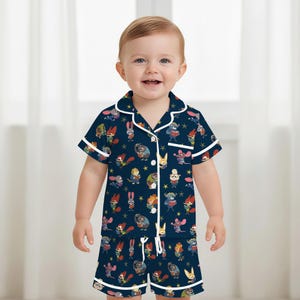 Pu&ograve; includere: Pigiama blu navy a maniche corte per bambini piccoli, con un motivo ripetuto di animali dei cartoni animati e stelle. Il set ha finiture e bottoni bianchi. I pantaloncini hanno un cordoncino. Il bambino sorride.