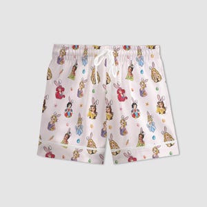 May include: White pajama shorts with an all-over print featuring cartoon princesses wearing bunny ears, carrots, and Easter eggs. The shorts have a drawstring waist and white trim.