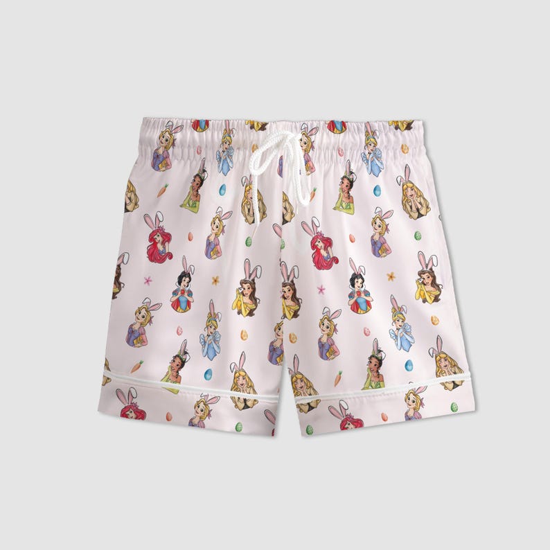 May include: White pajama shorts with an all-over print featuring cartoon princesses wearing bunny ears, carrots, and Easter eggs. The shorts have a drawstring waist and white trim.
