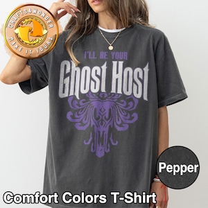 Puede incluir: Camiseta Comfort Colors gris oscuro con el texto blanco "I'LL BE YOUR Ghost Host" y un gráfico morado. La camiseta tiene cuello redondo y mangas cortas. La palabra "Pepper" está en un círculo en la parte inferior derecha.