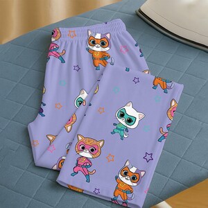 Puede incluir: Un pantal&oacute;n de pijama lavanda con un estampado repetido de gatos superh&eacute;roes de dibujos animados y formas de estrellas. Los gatos llevan m&aacute;scaras y disfraces en varios colores, como naranja, rosa y turquesa. El pijama est&aacute; doblado sobre una superficie azul.