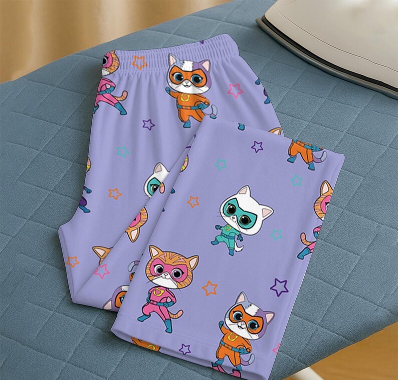 Puede incluir: Un pantal&oacute;n de pijama lavanda con un estampado repetido de gatos superh&eacute;roes de dibujos animados y formas de estrellas. Los gatos llevan m&aacute;scaras y disfraces en varios colores, como naranja, rosa y turquesa. El pijama est&aacute; doblado sobre una superficie azul.