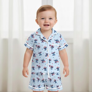 May include: A light blue pajama set for a toddler, featuring a repeating pattern of a cartoon character in patriotic attire. The short-sleeved top and shorts have white trim and buttons.