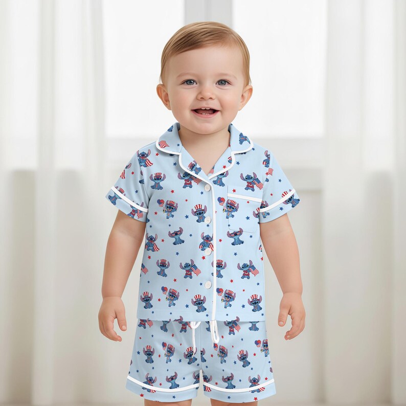 May include: A light blue pajama set for a toddler, featuring a repeating pattern of a cartoon character in patriotic attire. The short-sleeved top and shorts have white trim and buttons.