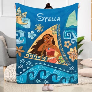 May include: A blue fleece blanket featuring a central illustration of the character Moana within a triangular frame. The design includes tropical flowers, ocean waves, and geometric patterns in shades of blue, orange, and yellow, with the name Stella printed at the top.