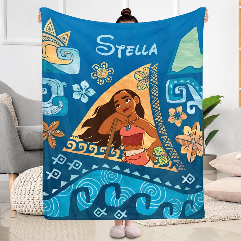 May include: A blue fleece blanket featuring a central illustration of the character Moana within a triangular frame. The design includes tropical flowers, ocean waves, and geometric patterns in shades of blue, orange, and yellow, with the name Stella printed at the top.