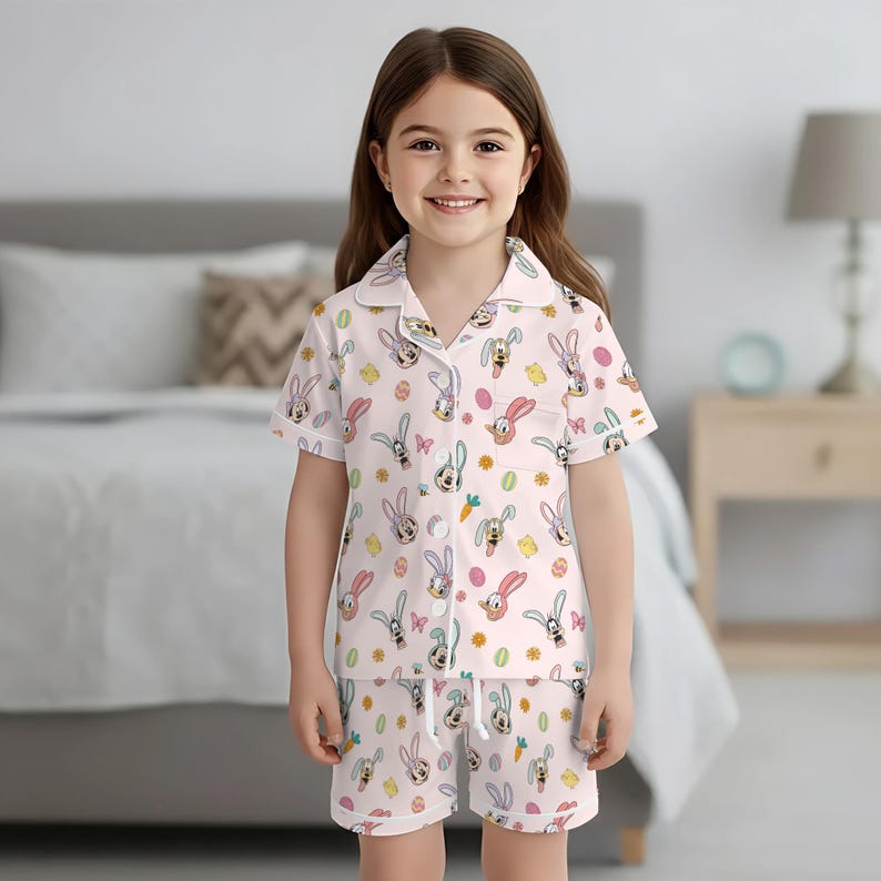 Puede incluir: Una ni&ntilde;a peque&ntilde;a con un conjunto de pijama rosa claro con un estampado repetido de conejitos de Pascua, huevos, zanahorias y flores. La parte superior de manga corta y los pantalones cortos tienen ribetes blancos. La ni&ntilde;a est&aacute; sonriendo.