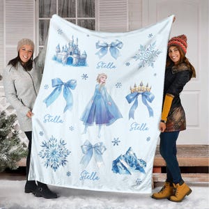 Op de afbeelding: Een lichtblauwe deken met illustraties van een kasteel, strikken, sneeuwvlokken, een kroon en een prinses. De naam "Stella" staat op de deken. De deken wordt door twee personen omhoog gehouden.