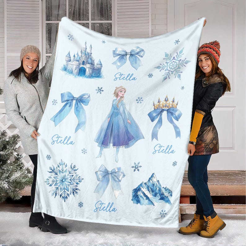 Op de afbeelding: Een lichtblauwe deken met illustraties van een kasteel, strikken, sneeuwvlokken, een kroon en een prinses. De naam "Stella" staat op de deken. De deken wordt door twee personen omhoog gehouden.