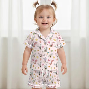 Puede incluir: Conjunto de pijama rosa claro con un estampado de conejitos de dibujos animados, huevos de Pascua y flores. La parte superior de manga corta y los pantalones cortos tienen ribetes blancos. El pijama est&aacute; hecho de una tela suave y ligera.