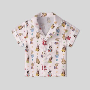 May include: White short-sleeved pajama top with a repeating pattern of cartoon princesses wearing bunny ears. The shirt has a collared neckline, a chest pocket, and button closures. The princesses are in various dresses and poses.