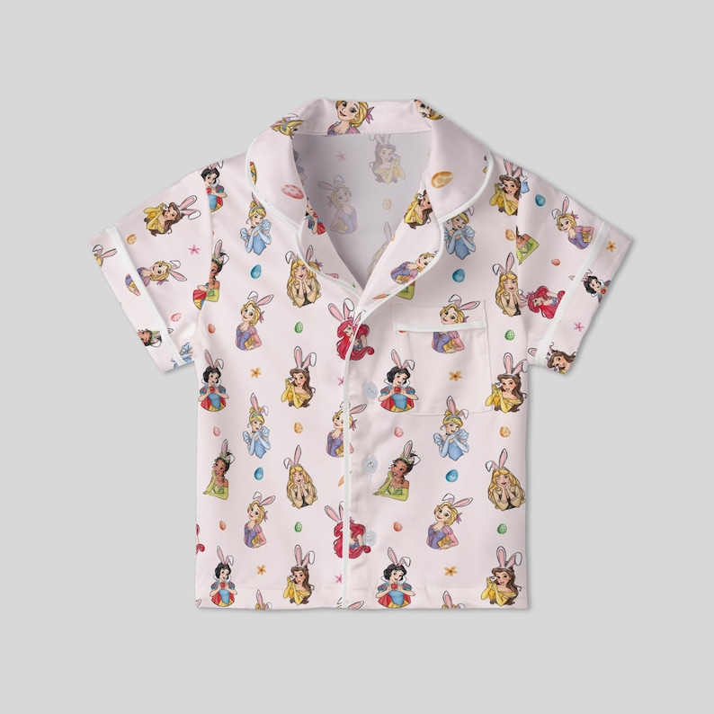 May include: White short-sleeved pajama top with a repeating pattern of cartoon princesses wearing bunny ears. The shirt has a collared neckline, a chest pocket, and button closures. The princesses are in various dresses and poses.