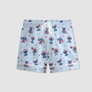 May include: Light blue pajama shorts with a repeating pattern of a blue cartoon character wearing patriotic accessories and holding American flags. The shorts have a white drawstring and white trim.