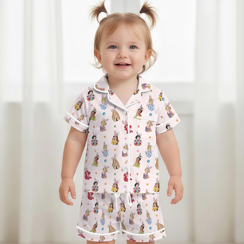 May include: A light pink pajama set for a toddler, featuring a short-sleeved button-up shirt and matching shorts. The set is adorned with a repeating pattern of cartoon bunnies in various colors and designs. White trim outlines the collar, cuffs, and edges.