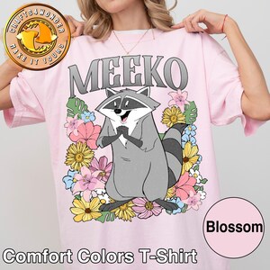 Pu&ograve; includere: T-shirt Comfort Colors rosa chiaro con un personaggio dei cartoni animati di procione di nome Meeko, circondato da fiori colorati. La parola "MEEKO" &egrave; stampata sopra il procione in lettere grigie. La maglietta ha anche la parola "Blossom" in un cerchio rosa.
