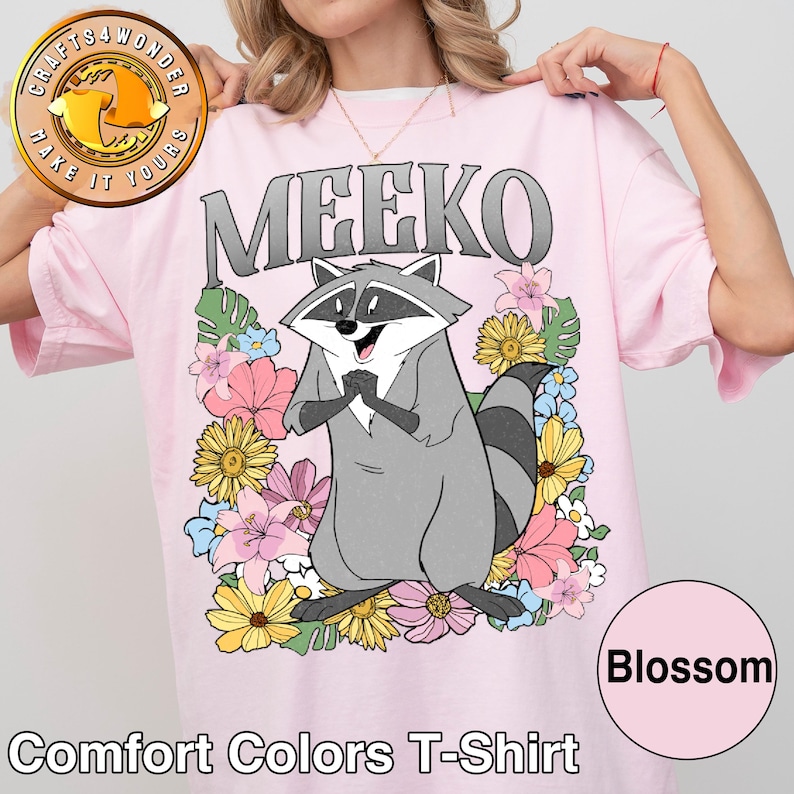Pu&ograve; includere: T-shirt Comfort Colors rosa chiaro con un personaggio dei cartoni animati di procione di nome Meeko, circondato da fiori colorati. La parola "MEEKO" &egrave; stampata sopra il procione in lettere grigie. La maglietta ha anche la parola "Blossom" in un cerchio rosa.
