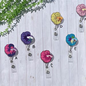 May include: A collection of six retractable badge reels featuring colorful betta fish designs. The fish have vibrant colors including pink, purple, yellow, and blue. Each reel has a clear plastic strap and a metal clip.