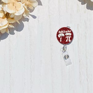 May include: A red glitter badge reel with the text "Math Teacher" and "Rational Get Real" with the symbols "i" and "π". The reel has a clear plastic clip and a silver metal button.