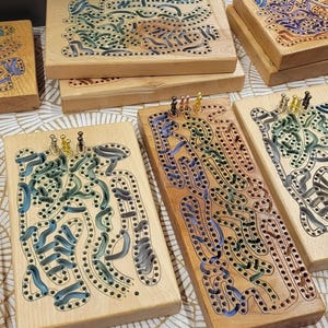 May include: A collection of wooden game boards with colourful, intricate patterns. The boards are made of light wood and have a variety of colours, including blue, green, red, and yellow. The boards are all different sizes and shapes, and they are all arranged in a row on a white surface.