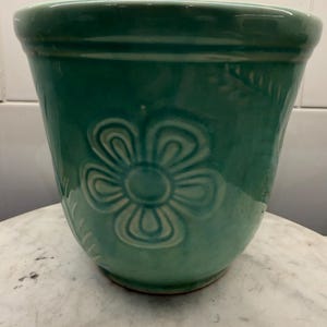 May include: A teal ceramic flower pot with a glossy finish and a raised, embossed flower design on the front. The pot features a wide, rounded rim and a tapered body, showcasing a consistent green-blue color throughout the glazed surface.
