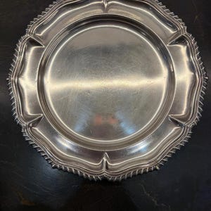 May include: A round silver-toned metal serving platter featuring a smooth, reflective central surface and a wide, scalloped rim with a detailed, textured rope-like edge pattern. The platter has a polished finish with visible light surface scratches.
