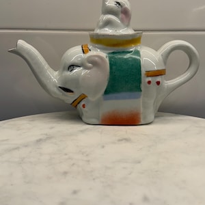 May include: A white ceramic teapot shaped like an elephant, with a matching elephant lid. The teapot features a green, yellow, and orange design. The spout and handle are curved. The teapot is sitting on a white marble surface.