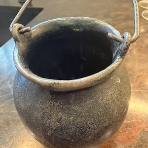 May include: A round, dark gray, textured pot with a wide opening and a thin, arched metal handle attached to two side loops. The exterior surface has a mottled, weathered appearance with a matte finish, and the interior is deep and dark.