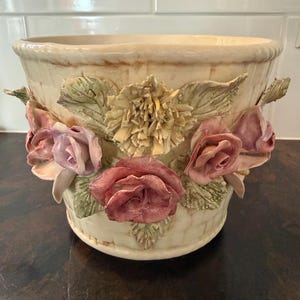 May include: A cream-colored ceramic planter featuring a textured basket-weave pattern on the exterior. The vessel is adorned with three-dimensional sculpted floral elements, including pink roses and a central pale yellow flower with green leaves.