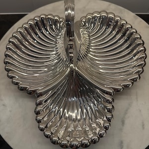 May include: A silver-toned metal serving dish shaped like a three-lobed shell with a central vertical handle. The surface features deep, radiating fluted grooves that extend from the center to the scalloped, rounded edges of each section.
