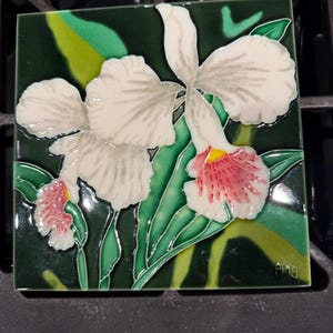May include: A square ceramic tile featuring a raised relief design of white orchid flowers with pink speckled centers and yellow accents, set against a dark green background with green leaves. The name Ping is signed in the bottom right corner.