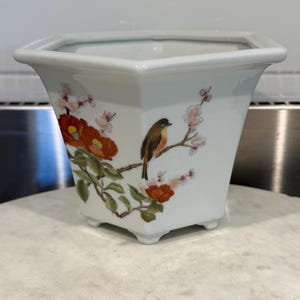 May include: A white hexagonal ceramic planter featuring a hand-painted design of a small brown and orange bird perched on a flowering branch with red and pink blossoms. The vessel has a flared rim and stands on four small, integrated feet.