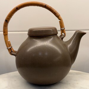 May include: A round, matte brown ceramic teapot featuring a smooth, spherical body, a short spout, and a matching lid. The teapot is equipped with a curved, light brown woven rattan handle that is attached to the body with metal loops.