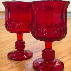 May include: Two matching ruby red glass goblets featuring a pressed diamond pattern around the midsection of the bowl and a textured, faceted stem design. The glasses have a wide, circular base and a flared rim, crafted from translucent red glass.