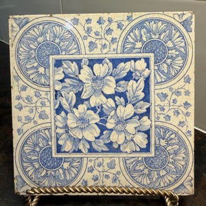 May include: A square ceramic tile featuring a blue and white floral pattern. The center displays a cluster of flowers within a square border, while each corner contains a circular motif with radiating petals and delicate vine accents throughout.
