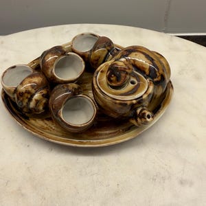 May include: A ceramic snail-shaped serving set featuring a brown and cream marbled glaze. The set includes a round tray, a lidded snail-shaped container, and three individual snail-shaped bowls with white interiors for serving food.