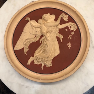 May include: A circular wall plaque featuring a cream-colored relief of a winged figure holding a small child against a deep terracotta red background. The piece is encased in a light beige wooden frame with a thin red accent line along the inner edge.