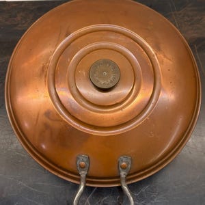 May include: A round copper lid with a central circular knob featuring the text "IMPORTANT PULL". The lid has a stepped design and a single U-shaped metal handle attached with two rivets at the bottom edge, showing a warm metallic finish.
