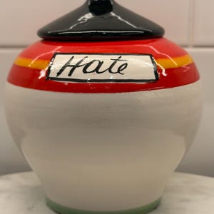 May include: A ceramic jar with a black lid and a red and white body. The jar has a yellow stripe and the word "Hate" written on it. The base of the jar is green.