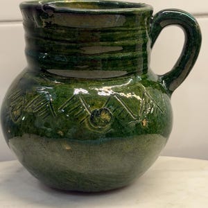May include: A round ceramic pitcher with a glossy dark green glaze and a visible crackle finish. The body features a series of incised triangular patterns around the center. It has a single curved handle on the right side and a wide opening.
