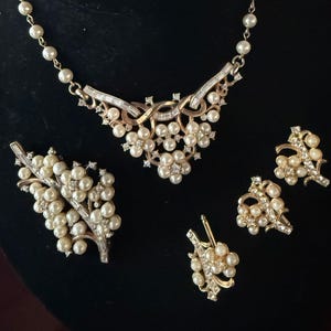 May include: A vintage jewelry set featuring a necklace, earrings, and a brooch. The set includes faux pearls and clear rhinestones set in gold-tone metal. The necklace has a floral design with a pearl strand. The earrings and brooch have a similar floral design.
