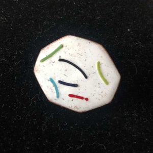 Könnte beinhalten: Ein weißer achteckiger Pin mit einer gesprenkelten Oberfläche und bunten Linien in Rot, Grün, Blau und Schwarz.