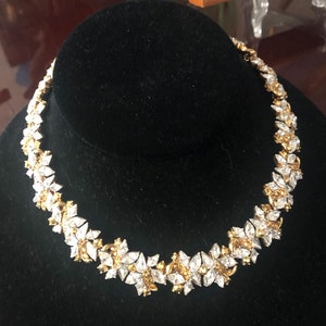May include: A gold and clear crystal necklace with a floral design. The necklace is on a black velvet stand.
