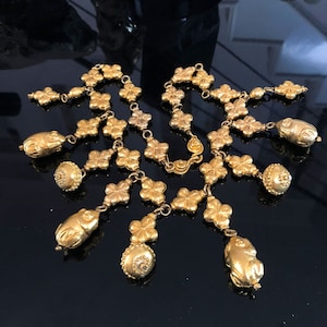 Op de afbeelding: Een gouden ketting met een ketting van bloemvormige schakels en hangende bedels. De bedels zijn rond en ovaal, sommige hebben een getextureerd oppervlak.