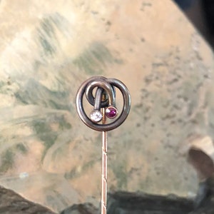 May include: A gold tie tack with a circular design featuring a small red stone and a smaller white stone.