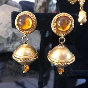 May include: A pair of gold tone drop earrings with amber colored glass stones. The earrings have a round top with a rope design and a smaller round stone dangling below.