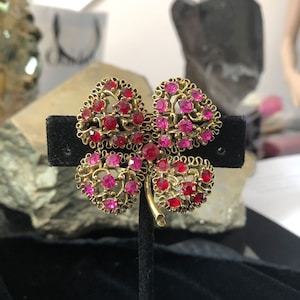 Puede incluir: Broche dorado con forma de trébol de cuatro hojas con pedrería roja y rosa. El broche tiene un diseño ornamental de filigrana y se exhibe sobre un soporte de terciopelo negro. Las hojas del trébol están llenas de piedras brillantes.