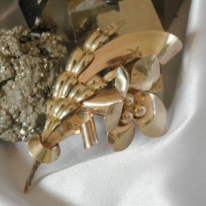 May include: A gold-toned metal brooch in the shape of a stylized flower with leaves. The flower has three petals and a center with three round accents.