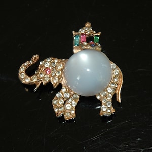 May include: A rhinestone-encrusted elephant brooch with a large, white cabochon stone and a crown on its head. The crown is decorated with colorful rhinestones.