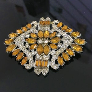 May include: A vintage brooch with a geometric design featuring a central cluster of amber-coloured rhinestones surrounded by smaller clear rhinestones.